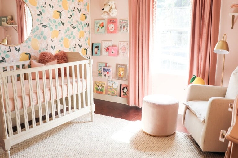 This image shows a baby's nursery decorated with pink and white hues. There is a crib with pink bedding, a white armchair, a round ottoman, and a wall adorned with patterned wallpaper and children's books display. Nursery themes come to life as natural light streams through the window framed by pink curtains.
