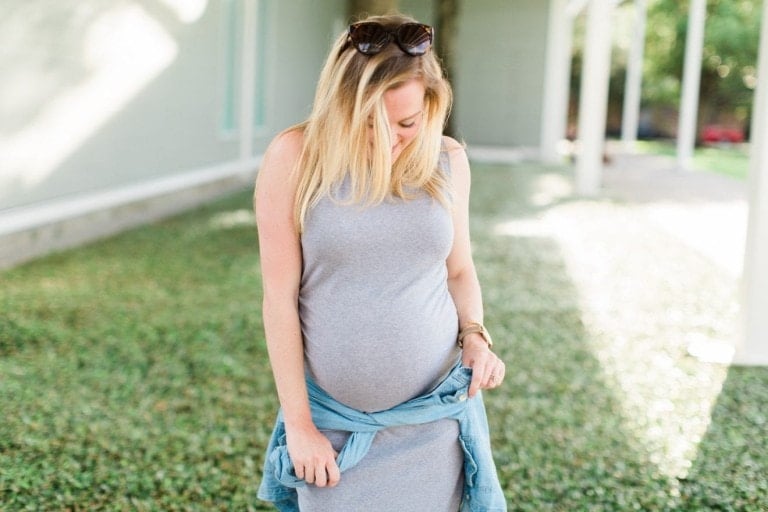 A woman with blonde hair wearing sunglasses on her head and a gray sleeveless dress stands outside. She has a light blue denim jacket tied around her waist and is looking down, one hand resting on her pregnant belly. The background shows a green lawn and a building, illustrating the perfect time to wear maternity clothes.