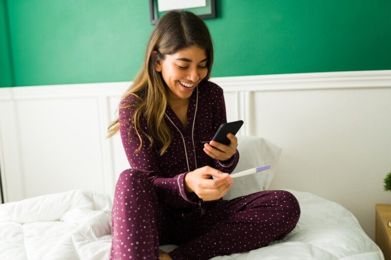 A woman wearing a maroon polka-dot pajama set sits cross-legged on a bed, looking at a smartphone in one hand and holding a pregnancy test in the other, with a smile on her face. The room has green walls with white wainscoting, reflecting the joy of early pregnancy.