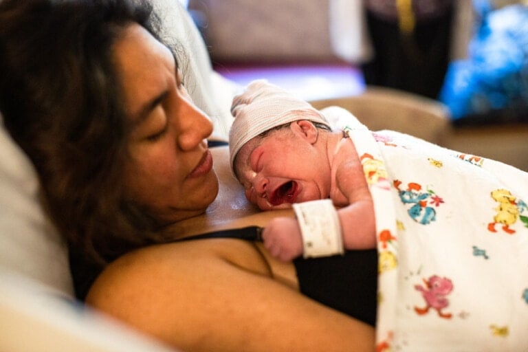 A newborn baby wrapped in a patterned blanket is lying on the chest of a person resting in a hospital bed after a vacuum birth. The baby wears a hat and an ID band while crying. The person, with long hair, appears to be resting.