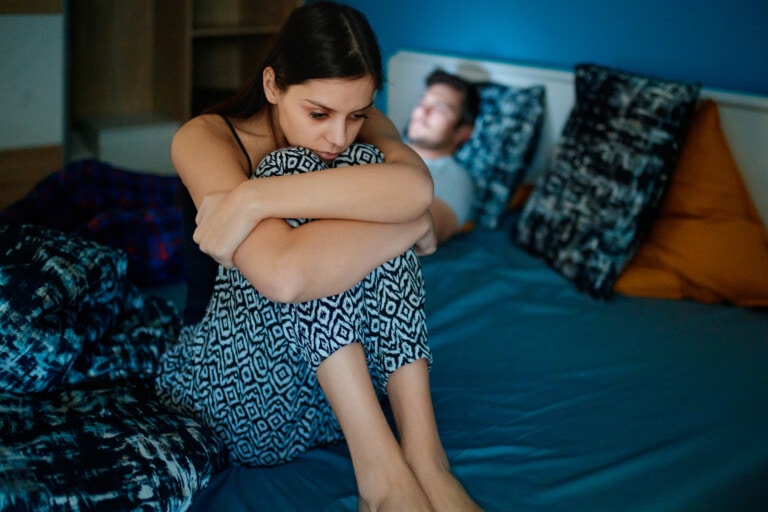 A woman sits on the edge of a bed with her knees drawn up to her chest, looking thoughtful. Clad in patterned pajamas, she is lost in contemplation. A man lies asleep in the background on the same bed, adding a touch of unspoken resentment toward your partner to the scene. The room has dim lighting.