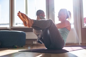 A woman is performing a seated yoga pose on an exercise mat while holding a baby. Balancing on her tailbone with her legs lifted and bent, she smiles at the baby. Natural light floods the room through a nearby window, creating an inspiring scene of fitness tips for new moms.